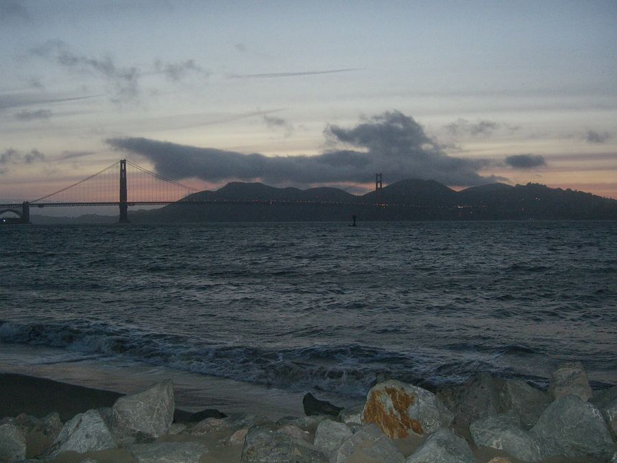 The Golden Gate Bridge silhouetted against the dusk sky across the choppy water of San Francisco Bay, with rocks on the shore in the foreground